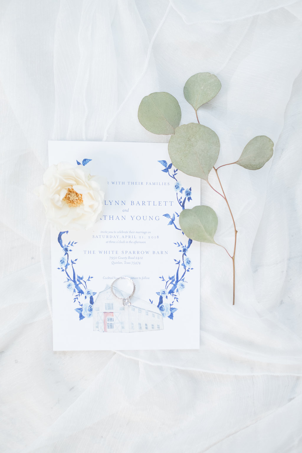 A professional image of the front of a formal wedding invitation card featuring blue and white flowers and barn painting Motif.
